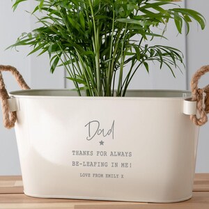 May include: White metal planter with rope handles, featuring the text "Dad Thanks for always be-leafing in me! Love from Emily x". A green plant is inside the planter.