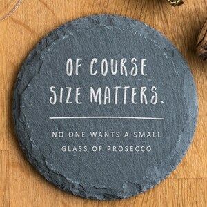 May include: A round, grey slate coaster with the text "OF COURSE SIZE MATTERS. NO ONE WANTS A SMALL GLASS OF PROSECCO" in white lettering. A champagne cork and wire cage sit nearby on a wooden surface. A wine glass is visible.