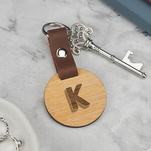 May include: A wooden keyring featuring the letter "K" on a circular, light-coloured wooden disc. It has a brown leather strap and a silver key-shaped bottle opener. Rings are visible in the background.