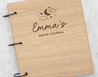May include: A light brown wooden journal with a natural grain finish, bound by three black metal rings on the left side. The cover features an engraved design of a crescent moon, clouds, and stars above the text Emma's DREAM JOURNAL in black.