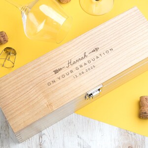 May include: A light-colored wooden wine box with the text "Hannah" and "ON YOUR GRADUATION 13.08.2025" engraved on the lid. The box is rectangular with a metal clasp. Wine glasses and corks are scattered around the box on a yellow surface.
