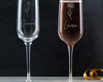 May include: Two personalized champagne flutes with floral designs and names "Gemma" and "Lauren" etched on the glass. The flutes are on white coasters.