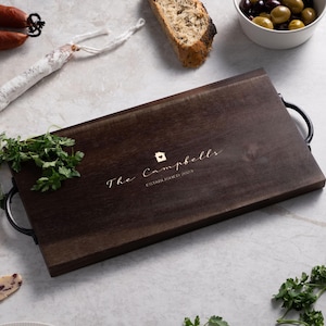 May include: A rectangular dark brown wooden serving tray with black metal handles on each side. The centre of the tray features a small house icon with a heart and the engraved text "The Campbells ESTABLISHED 2023" in a script font.