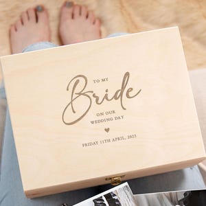 May include: A wooden box with a hinged lid and a brass latch. The lid is engraved with the text "To my Bride on our Wedding Day Friday 11th April, 2025" and a small heart.