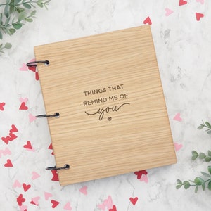 May include: A rectangular light brown wooden scrapbook with three black metal binder rings on the left side. The front cover features the engraved text "THINGS THAT REMIND ME OF you" with a small heart symbol centred below the text.