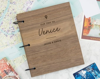 May include: A wooden photo album with a brown woodgrain finish. The cover is engraved with the text "Our Trip to Venice" and "Jacob & Annie".