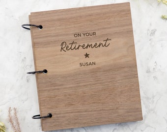 May include: A rectangular wooden scrapbook with a natural brown wood grain finish. The cover is engraved with the text ON YOUR Retirement, a small star symbol, and the name SUSAN. Three black metal binder rings secure the wooden cover on the left side.