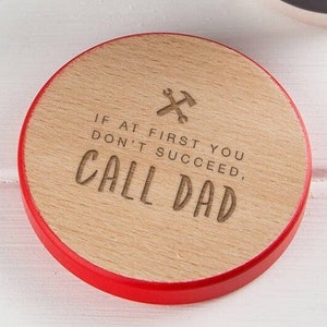 Peut inclure: Un dessous de verre rond en bois avec un bord rouge. Le dessous de verre porte l'inscription "IF AT FIRST YOU DON'T SUCCEED, CALL DAD" et un motif de marteau et de clé croisés.