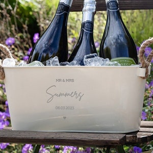May include: White metal ice bucket with rope handles, personalized with "MR & MRS Summers 06.03.2023". Three bottles of champagne are inside the bucket, surrounded by ice. Two champagne flutes are on a gold tray.