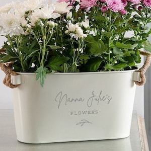 May include: A white oval metal planter with rope handles, filled with white and pink chrysanthemums. The planter has the text "Nanna Julie's Flowers" printed on the front.