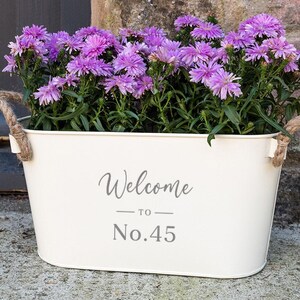 May include: White metal planter with rope handles, filled with purple flowers. The planter has the text "Welcome to No. 45" printed on the front.