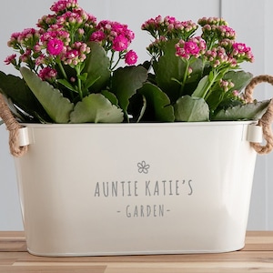 May include: White metal planter with rope handles, filled with pink flowers and the text "Auntie Katie's - GARDEN -"