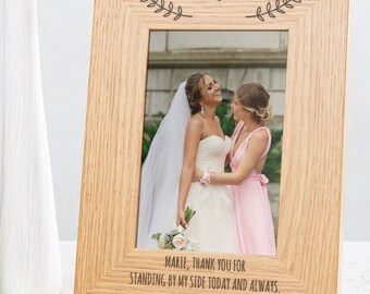 May include: A wooden picture frame with the word "Bridesmaid" engraved on the top. The frame has a photo of two women, one in a white wedding dress and the other in a pink bridesmaid dress. The text below the photo reads "Marie, thank you for standing by my side today and always. Love, Samantha."