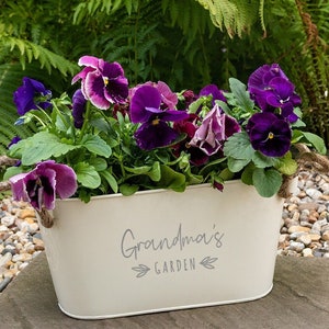 May include: White metal planter with rope handles, filled with purple and white pansies. The planter has the text "Grandma's Garden" printed on the front.