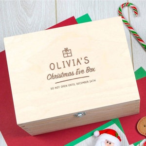 Puede incluir: Una caja de madera con tapa abatible y cierre. La caja est&aacute; personalizada con el texto "Olivia's Christmas Eve Box" y "No abrir hasta el 24 de diciembre".