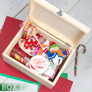 Puede incluir: Una caja de madera llena de art&iacute;culos navide&ntilde;os, incluyendo un juguete de mu&ntilde;eco de nieve, bastones de caramelo, un copo de nieve de fieltro y un adorno de huella de mano. La caja est&aacute; abierta y los art&iacute;culos son visibles en el interior. La caja est&aacute; etiquetada como "BOX 1".