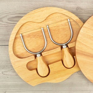 May include: A wooden cheese board with two metal cheese knives. The board has a round shape with a scalloped edge. The knives have wooden handles and are designed for cutting cheese.