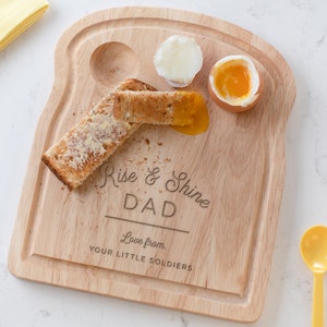 May include: Wooden breakfast serving tray shaped like a slice of toast with two egg cups and a space for toast. The tray has the text "Rise & Shine Dad Love from. Your Little Soldiers" engraved on it.