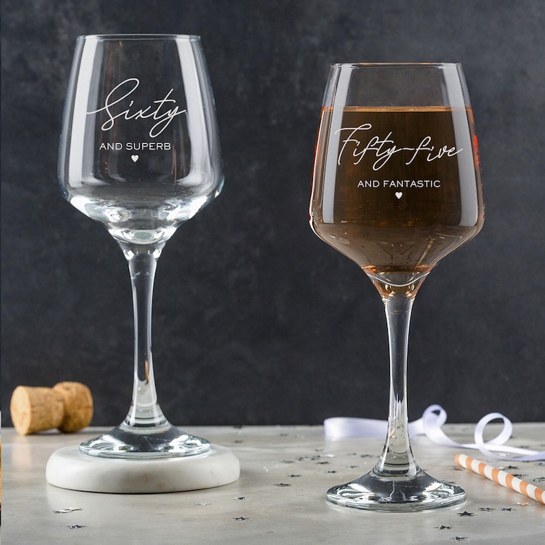 Puede incluir: Dos copas de vino de vidrio transparente con texto grabado. La copa de la izquierda dice "Sesenta y S&uacute;per" y la copa de la derecha dice "Cincuenta y cinco y Fant&aacute;stico".