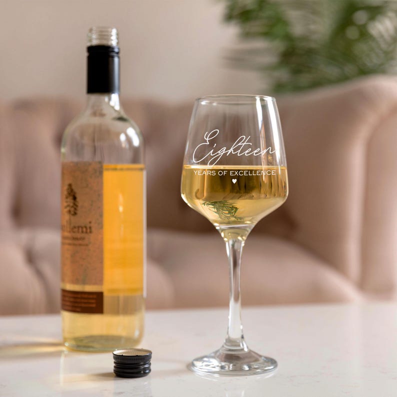 Puede incluir: Una copa de vino transparente con el texto "Eighteen Years of Excellence" grabado en el lateral. La copa est&aacute; llena de vino blanco y est&aacute; sobre una mesa blanca junto a una botella de vino.
