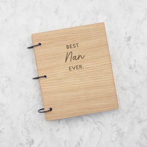 May include: A rectangular wooden scrapbook with a light brown wood grain finish. The front cover features the engraved text "BEST Nan EVER." in a combination of block and script fonts. Three black metal binder rings secure the cover on the left side.