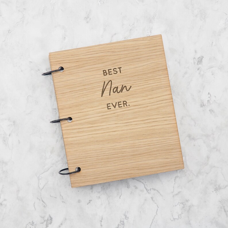 May include: A rectangular wooden scrapbook with a light brown wood grain finish. The front cover features the engraved text "BEST Nan EVER." in a combination of block and script fonts. Three black metal binder rings secure the cover on the left side.