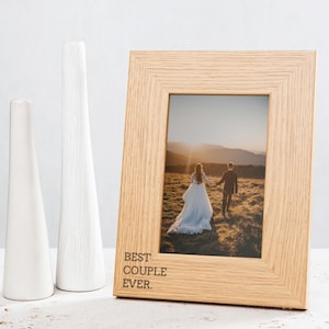 May include: A wooden photo frame with a light brown finish. The frame has a white mount and a photo of a couple walking in a field. The frame has the text "BEST COUPLE EVER." engraved on the bottom.