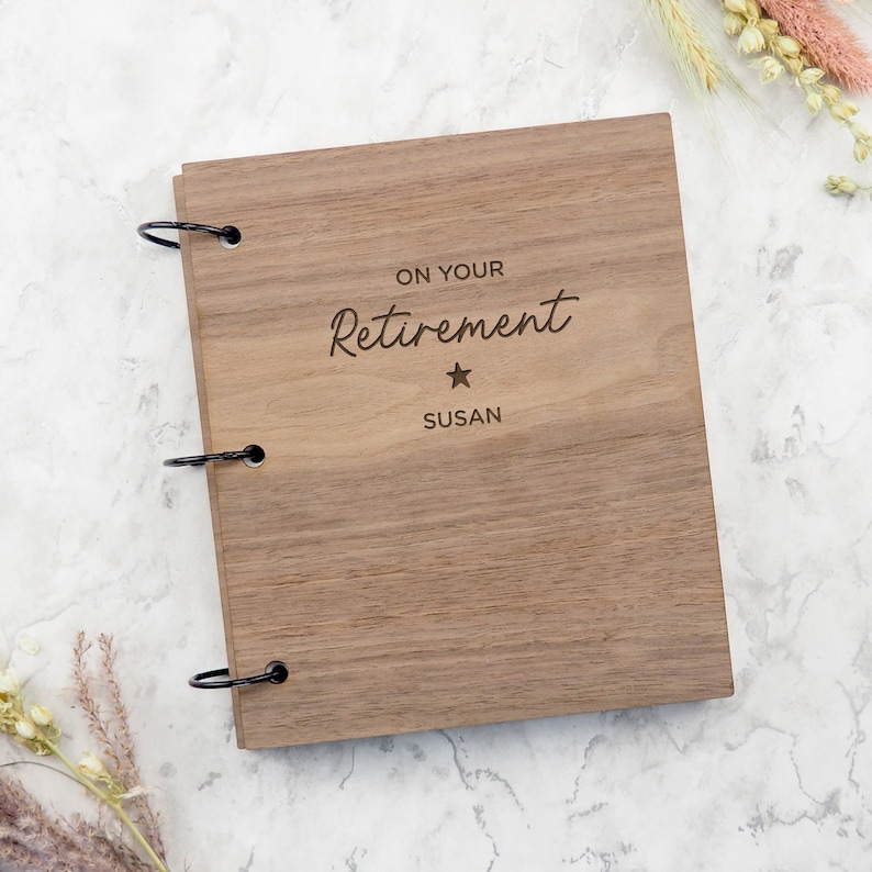 Puede incluir: Un &aacute;lbum de recortes rectangular de madera con un acabado de veta de madera marr&oacute;n natural. La cubierta est&aacute; grabada con el texto ON YOUR Retirement, un peque&ntilde;o s&iacute;mbolo de estrella y el nombre SUSAN. Tres anillas met&aacute;licas negras sujetan la tapa.