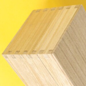 May include: Close-up of a wooden box with dovetail joints. The box is made of light-colored wood, with visible grain patterns. The box is set against a bright yellow background.