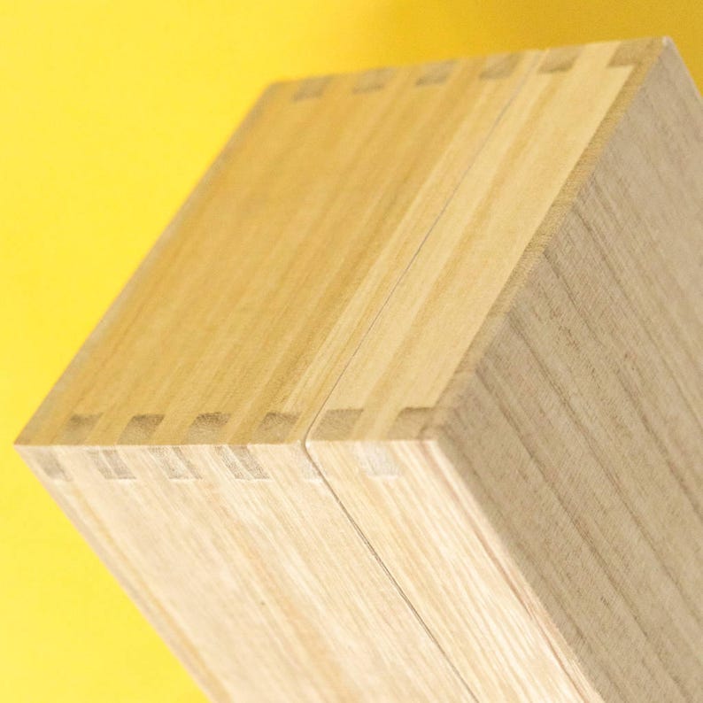 May include: Close-up of a wooden box with dovetail joints. The box is made of light-colored wood, with visible grain patterns. The box is set against a bright yellow background.