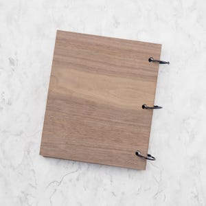 May include: A rectangular wooden cover with a natural brown wood grain pattern. The cover features three circular holes along the right edge, each fitted with a black metal binder ring to secure the pages together in a vertical orientation.
