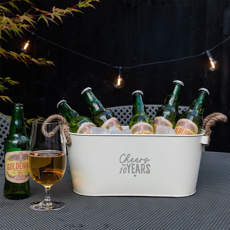 May include: An oval white metal beverage tub with thick rope handles, featuring the text "Cheers to 10 YEARS" with a small heart icon. The tub is filled with ice and five green glass beer bottles, with one additional bottle and a glass of beer nearby.