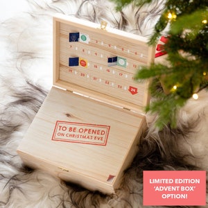 Puede incluir: Una caja de calendario de adviento de madera con 24 puertas numeradas. La caja est&aacute; abierta y las puertas son visibles. La caja tiene un sello rojo que dice "Para abrir en Nochebuena".