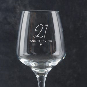Puede incluir: Una copa de vino de vidrio transparente con el texto "21 and thriving" y un peque&ntilde;o s&iacute;mbolo de coraz&oacute;n.