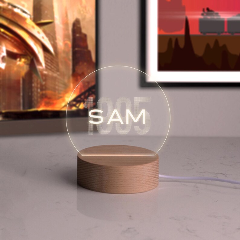 May include: A personalized LED night light with the name "SAM" and the number "1995" etched into a clear, circular acrylic panel. The panel is illuminated by a warm white light and sits on a round wooden base.