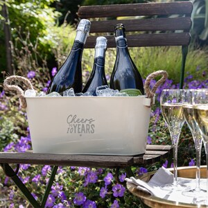 May include: A cream-colored metal beverage tub with two jute rope handles, featuring the text "Cheers to 10 YEARS" in gray. The tub is filled with ice and three dark glass bottles of sparkling wine, accompanied by two clear glass flutes nearby.