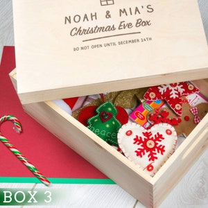 Puede incluir: Una caja de madera con una tapa que dice "NOAH & MIA'S Christmas Eve Box" y "DO NOT OPEN UNTIL DECEMBER 24TH". La caja est&aacute; abierta y contiene una variedad de adornos navide&ntilde;os, incluyendo un coraz&oacute;n de fieltro con un copo de nieve, un hombre de jengibre de fieltro y un &aacute;rbol de Navidad de fieltro.