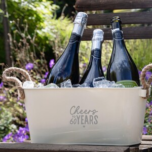 May include: A cream-colored metal beverage tub with two jute rope handles, filled with ice and three dark glass bottles of Brut sparkling wine. The front of the tub features the gray printed text Cheers to 60 YEARS with a small heart icon.