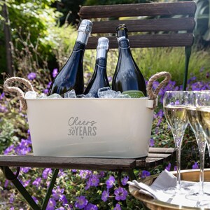 May include: A cream-colored metal beverage tub with two rope handles, filled with ice and three dark glass bottles. The front of the tub features the text Cheers to 30 YEARS in a dark gray script and block font, with a small heart icon below.