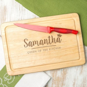 May include: A personalised wooden chopping board with the text "Samantha Queen of the Kitchen" and a crown design. A red kitchen knife is placed on the board.