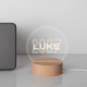 May include: A personalized LED night light with a wooden base. The clear acrylic light features the name "LUKE" and the year "2007" in a white font, illuminated by a soft glow. The light is connected to a white cord.
