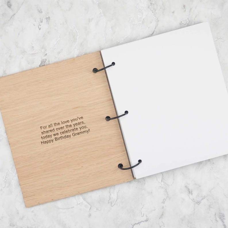 May include: A light-coloured wooden scrapbook with three black metal rings holding blank white pages. The front cover features the engraved text: For all the love you've shared over the years, today we celebrate you. Happy Birthday Grammy!
