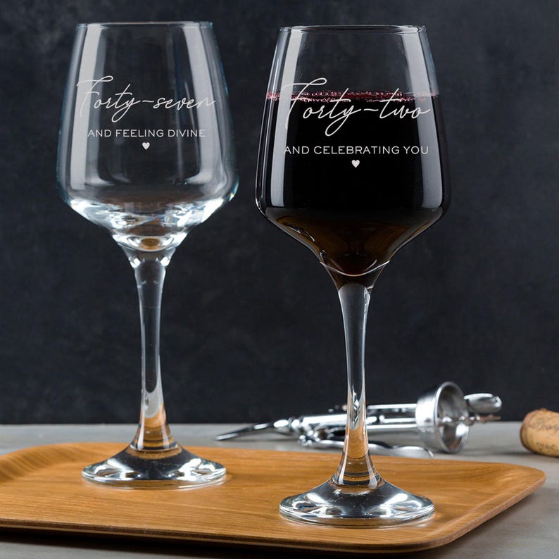 Puede incluir: Dos copas de vino transparentes con el texto "Forty-seven and feeling divine" y "Forty-two and celebrating you" grabado en ellas. La copa de la derecha est&aacute; llena de vino tinto.