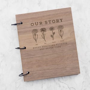 Puede incluir: Un álbum de recortes rectangular de madera con tres anillas de metal negro en el lado izquierdo. La cubierta presenta el texto grabado OUR STORY, cuatro ilustraciones florales de línea con los nombres Mum, Dad, Jacob y Sarah, y la frase WHERE MEMORIES BLOSSOM AND NEVER FADE.