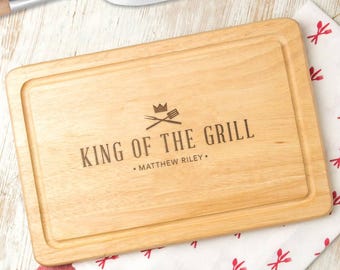 May include: A rectangular light-coloured wooden cutting board with a juice groove along the perimeter. The centre features an engraved crown icon, crossed grilling tools, and the text "KING OF THE GRILL" followed by "MATTHEW RILEY".