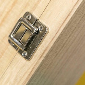 May include: Close-up of a silver metal latch with a rectangular clasp, attached to a light-colored wooden surface with visible wood grain. The latch is secured with four small screws.