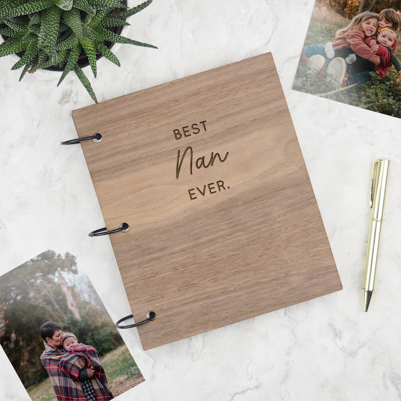 May include: A rectangular wooden photo album with a natural brown wood grain finish. The cover is engraved with the text BEST Nan EVER. The album is bound with three black metal rings on the left side. A gold pen is positioned to the right of the album.