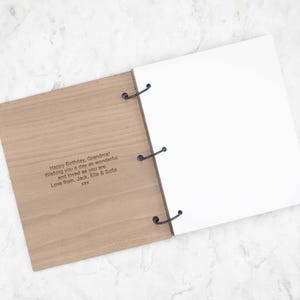 May include: A wooden scrapbook with three black metal rings holding blank white pages. The front wooden cover features the engraved text: Happy Birthday, Grandma! Wishing you a day as wonderful and loved as you are. Love from, Jack, Elle & Sofia xxx.