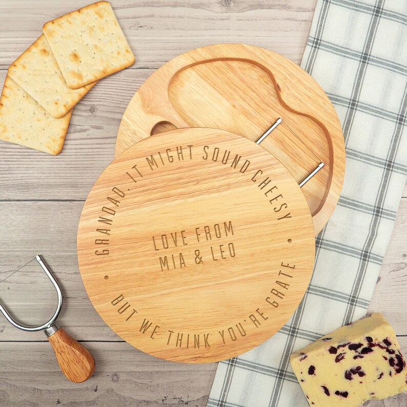 May include: A round wooden cheese board with a built-in knife and a small well for crackers. The board is engraved with the text "Grandad. It might sound cheesy. But we think you're grate. Love from Mia & Leo."