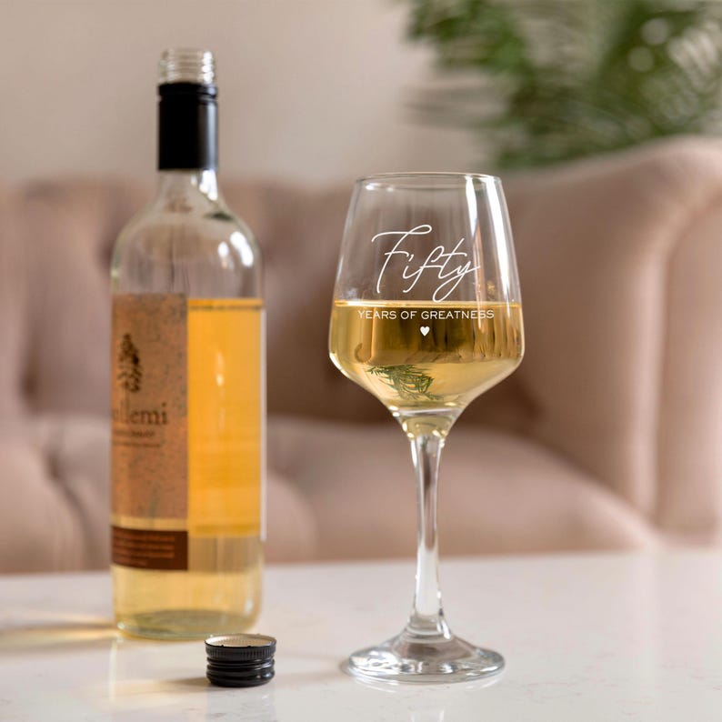 Puede incluir: Una copa de vino transparente con el texto "Fifty Years of Greatness" grabado en el lateral. La copa est&aacute; llena de vino blanco y est&aacute; sobre una mesa junto a una botella de vino.